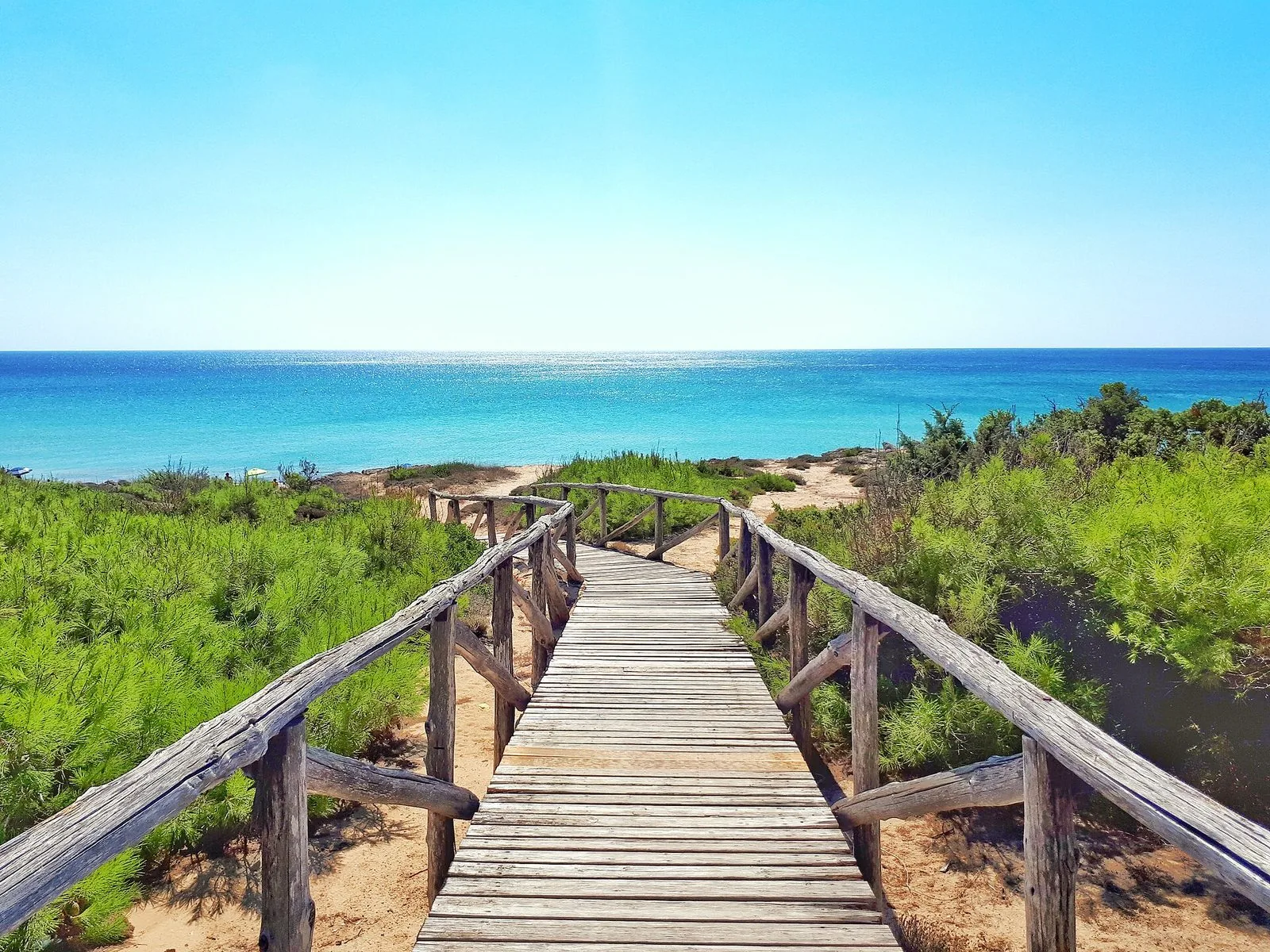 Passerella in legno tra le dune coperte di macchia mediterranea che scende verso il mare turchese a Campomarino di Maruggio