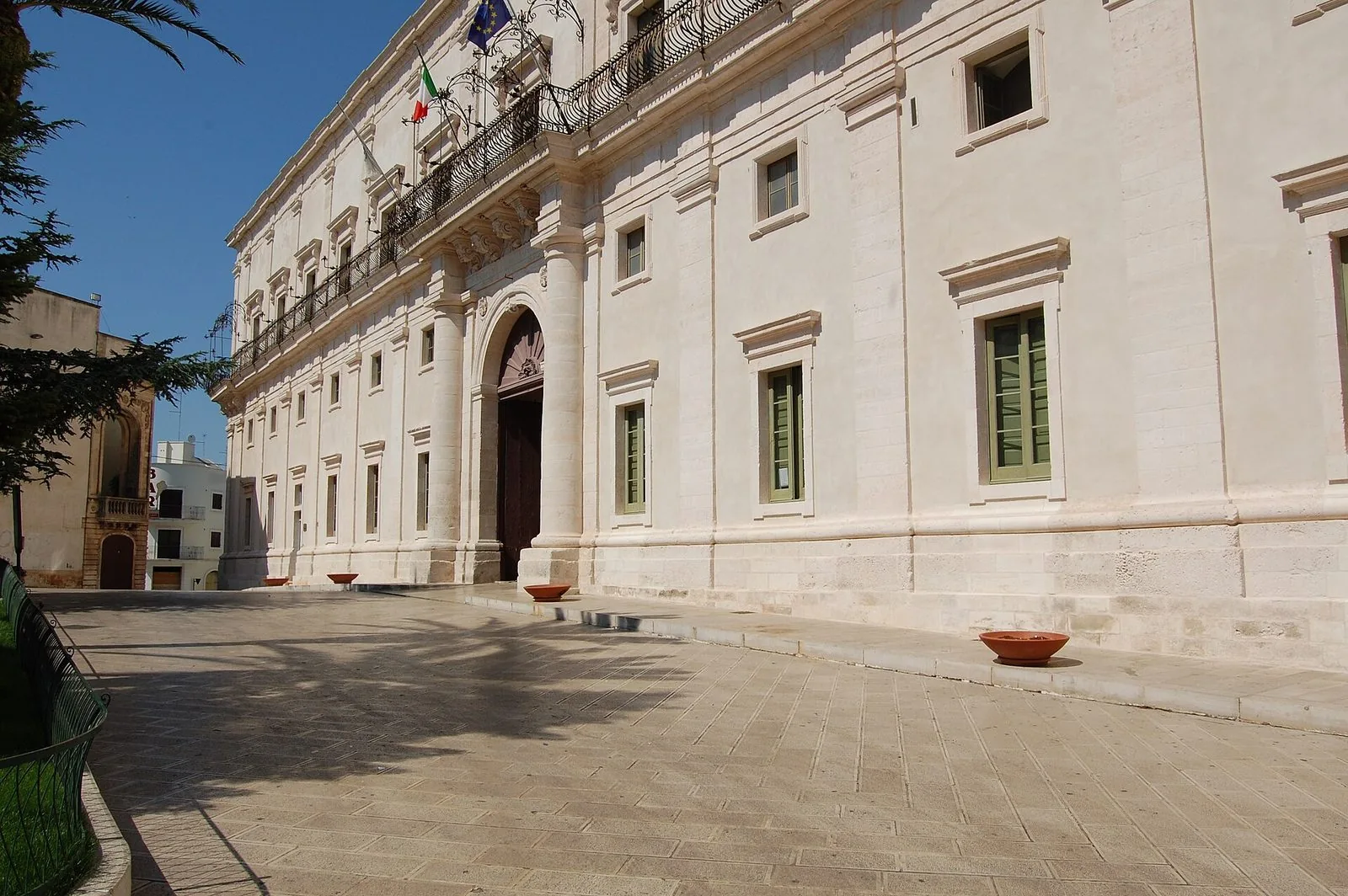Facciata chiara di Palazzo Ducale a Martina Franca con portale ad arco, balcone in ferro battuto e bandiere, in una giornata di sole