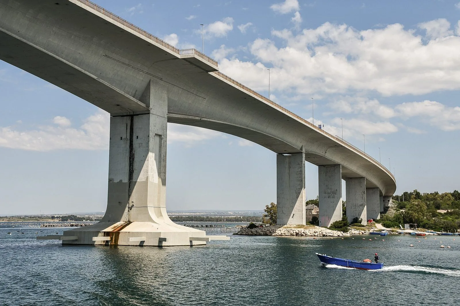 Il Ponte Punta Penna Pizzone visto dal basso sul Mar Piccolo di Taranto, con i piloni in cemento e una barca blu che passa sull'acqua